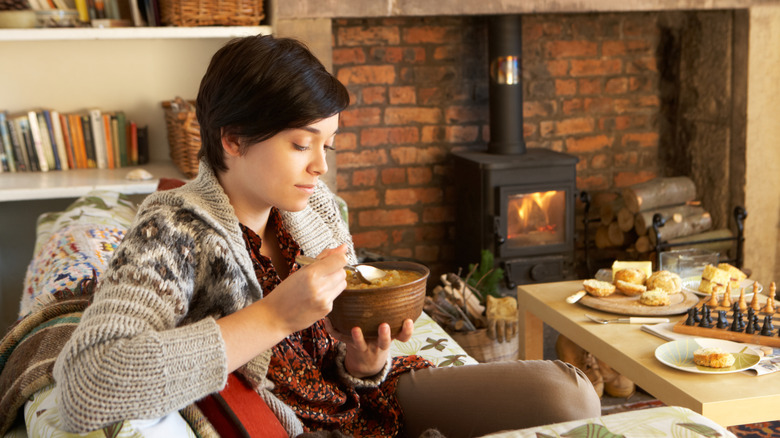 Young woman eating soup indoors sitting by fire