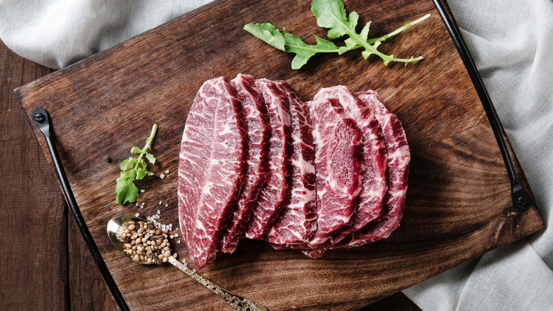 sliced raw beef brisket rests on a wooden cutting board, herbs surrounding it.