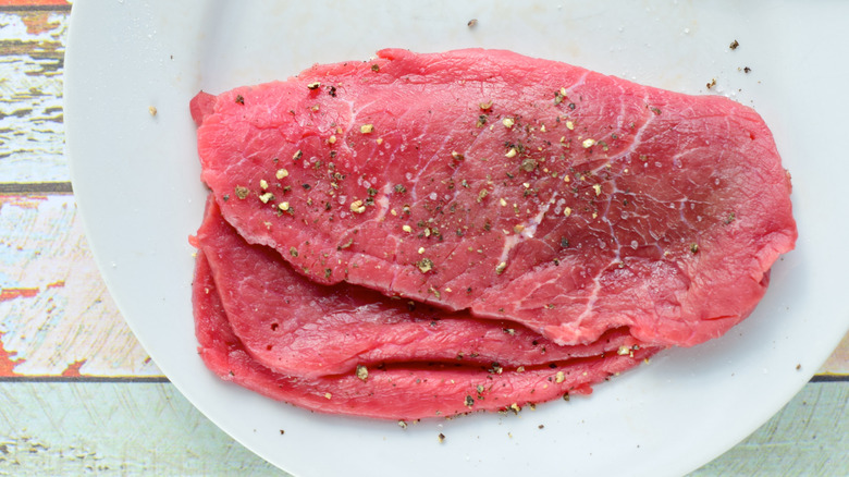 A stack of raw minute steaks on a white plate, seasonings sprinkled on top.