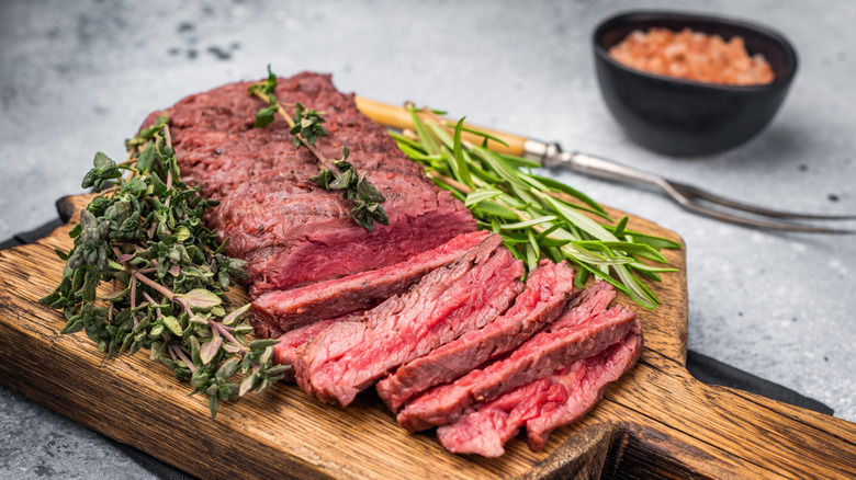 Grilled flank steak sliced into thin pieces rests on a wooden cutting board, herbs surrounding it.