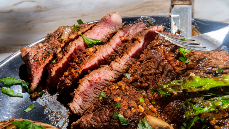 A cooked sirloin tip steak, sliced into thin pieces. Various greens line the plate, with a fork resting on the meat.