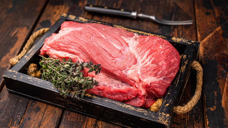 A cut of top round beef rests inside of a wooden tray, rosemary perched on the side.