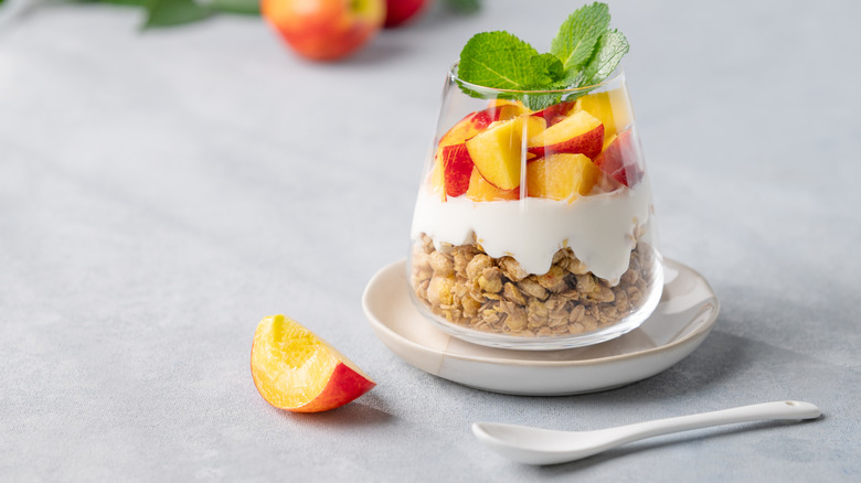 A yogurt parfait with peach and granola in a glass on a light background with fresh fruits
