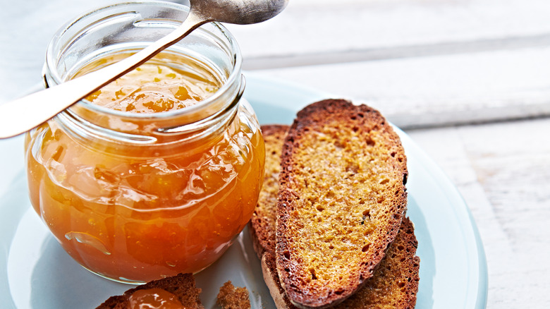 A jar of peach jam next to slices of toasted bread on a neutral background
