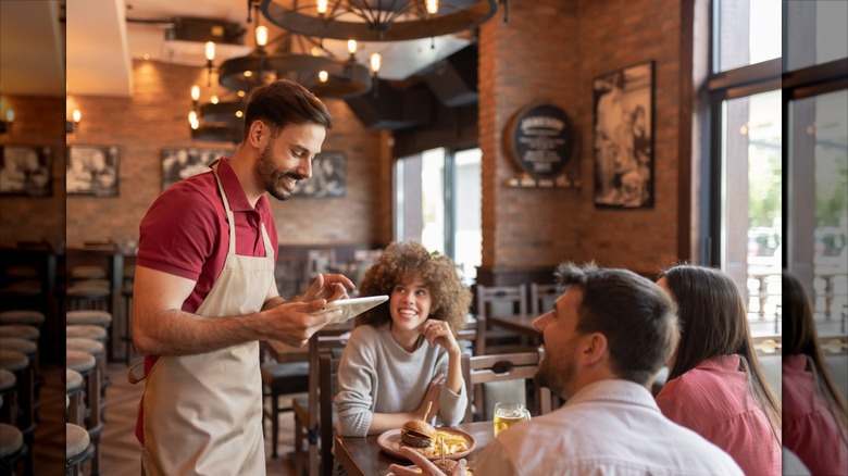 Waiter standing near guests while entering order on a tablet in a rustic-style bar