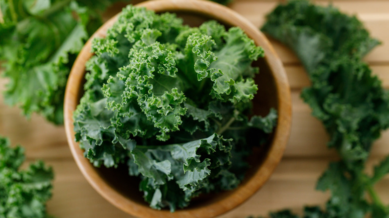 Kale leaves sit inside a wooden bowl. More leaves rest to the right and left of the bowl.
