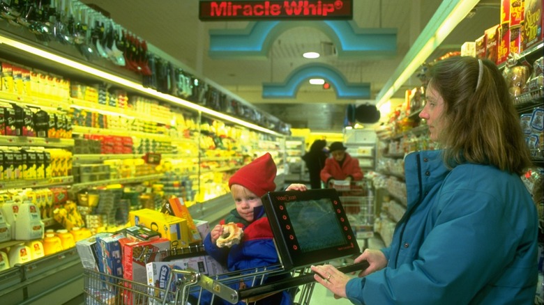 Mother and son in '90s supermarket aisle with shopping cart full of snacks