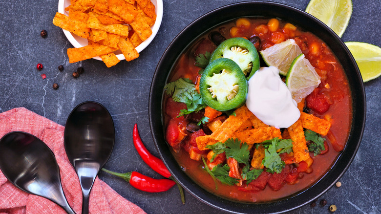 A stylized overhead shot of a bowl of tortilla soup topped with tortilla strips, sour cream, cilantro, lime, and jalapeño