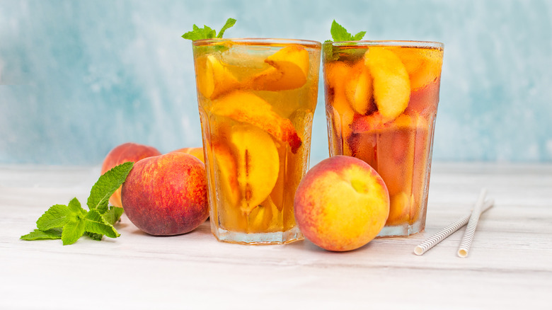 Peach ice tea in a glass with mint and whole peaches on white wooden table