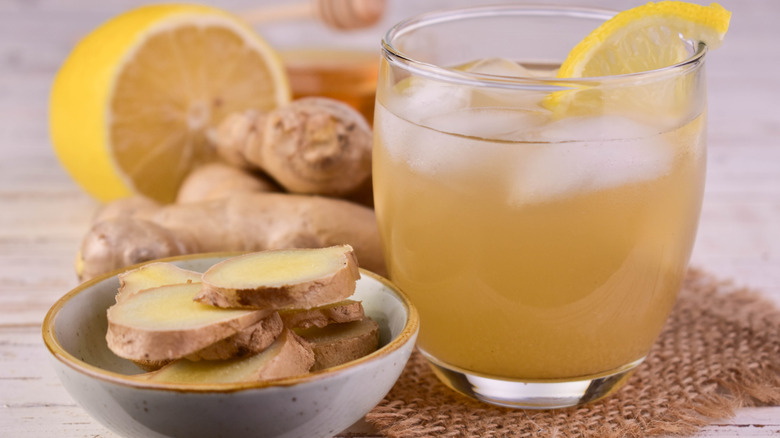 Ginger tea with ice, honey, and lemon in a glass perched on burlap surrounded by slices of fresh ginger in a bowl and