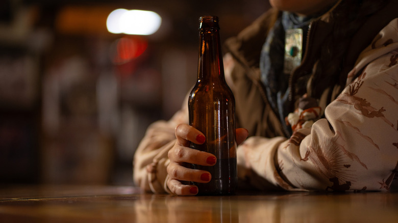 A woman dressed in rustic attire sitting at a bar and holding an open beer bottle.