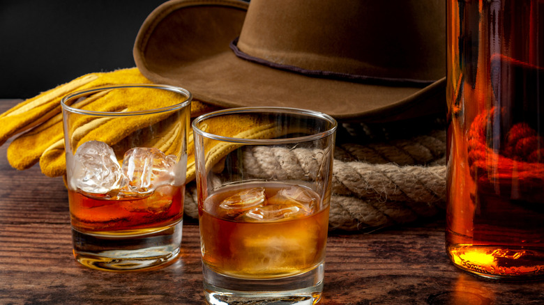 Two glasses of whiskey on the rocks, a bottle to the right, a cowboy hat, rope and gloves in the background resting on the bar.