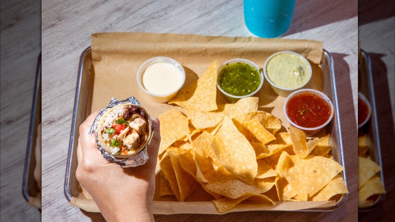 Hand holding Moe's burrito with chips and salsa on tray