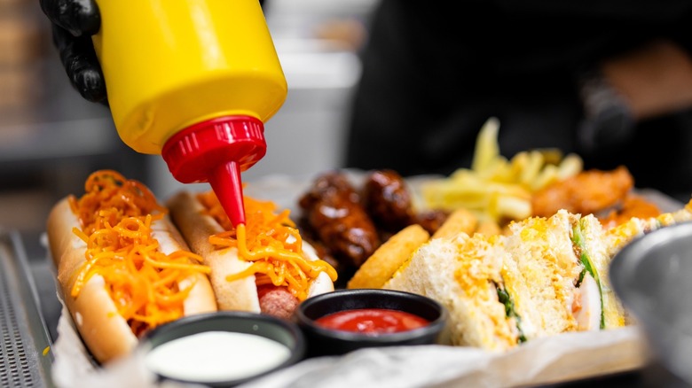 A chef in a black glove squirts yellow mustard onto a loaded hot dog on a platter with another hot dog, fries, wings, onion rings, and a sandwich, emphasizing fast food