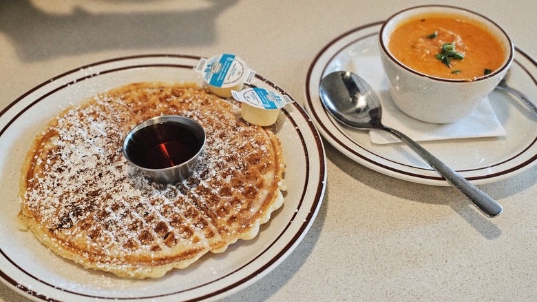 A plate of waffle with maple syrup and butter beside a bowl of tomato soup at Waffle House Diner