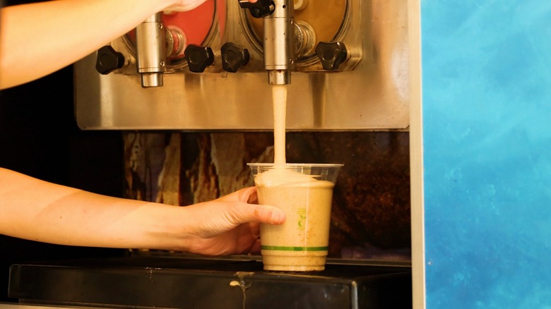 An employee at fast food restaurant uses a commercial machine to pour a cold frozen slushy into a plastic cup