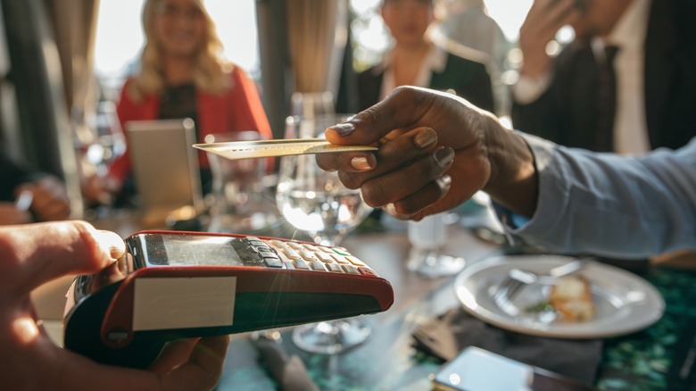close up of dinner party in background and close up of hand extending a card to a waiter to pay with a card reader