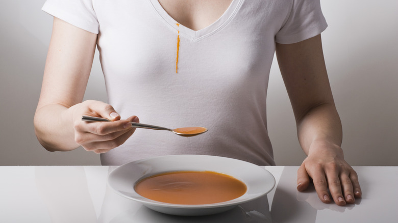 Woman with bowl of tomato soup on table in front of her, with soup spill stain on her white shirt