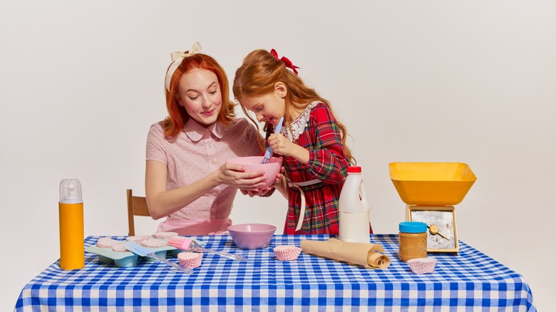 Portrait of mother and daughter cooking cake together isolated over grey background. Spending time together. Concept of beauty, retro style, fashion, elegance, 60s