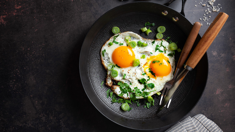 Two fried eggs in a skillet garnished with green onions and herbs with a fork and knife resting against the edge of the pan