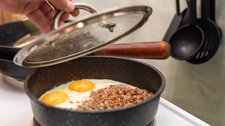 Someone removing the lid from a frying pan containing two eggs and some sausage