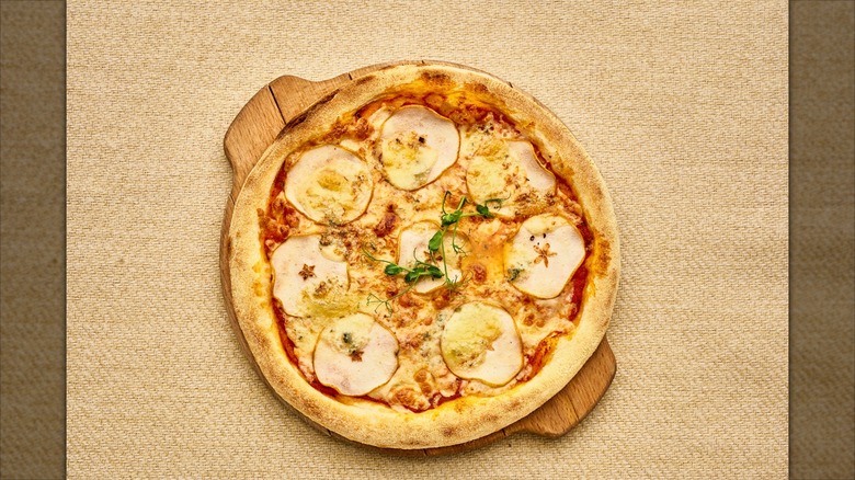 A pizza with a wooden tray on a table. The pizza has a lot of cheese and a few slices of apples