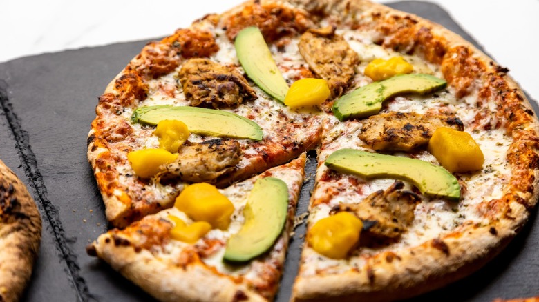 A closeup shot of a round pizza topped with avocado, chicken and mangos