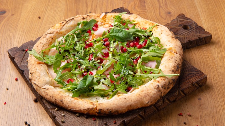 Oven-cooked Italian pizza Melagrano with Mozzarella cheese, ham, beef, arugula, pomegranate seeds, basil and olive oil. Hot crispy pizza on a wooden board.