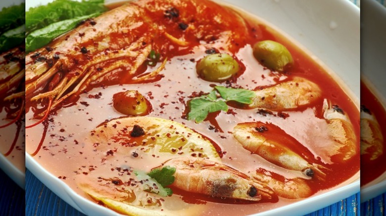 bowl of soup with reddish broth filled with seafood like shrimp and olives