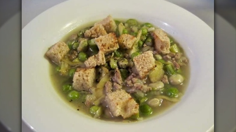 bowl of soup with green colored broth, fava beans, peas, and other green vegetables, topped with croutons