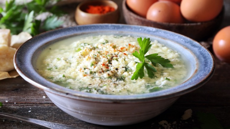 bowl of Italian egg drop soup with herb garnish sits on a table with other ingredients like eggs, cheese, and spices