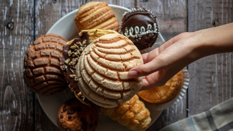 Hand shown holding a Concha over a plate filled with assorted Mexican pastries and breads
