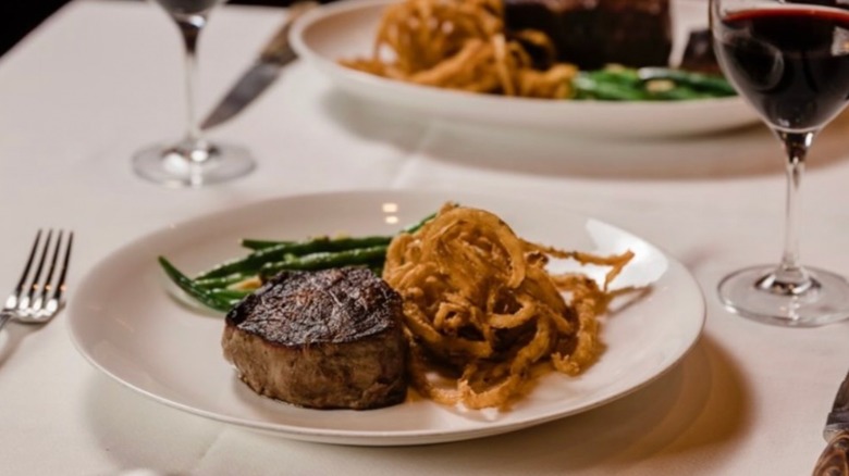 Filet Mignon with green beans and fried onions on a white plate, and a glass of red wine, all on a white tablecloth at Bern's Steak House