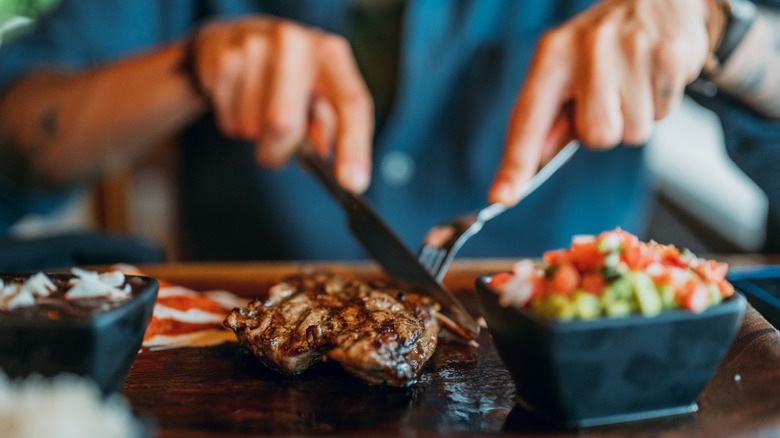 Hands holding fork and knife and eating delicious juicy steak on a wooden plate that is accompanied by a salad in a black square bowl