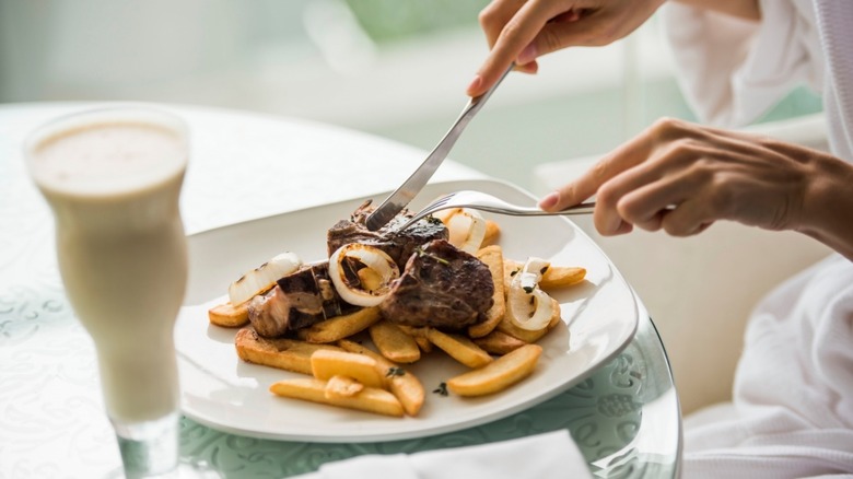 Hands shown cutting into a steak on a white [late with a side of French fries at a restaurant