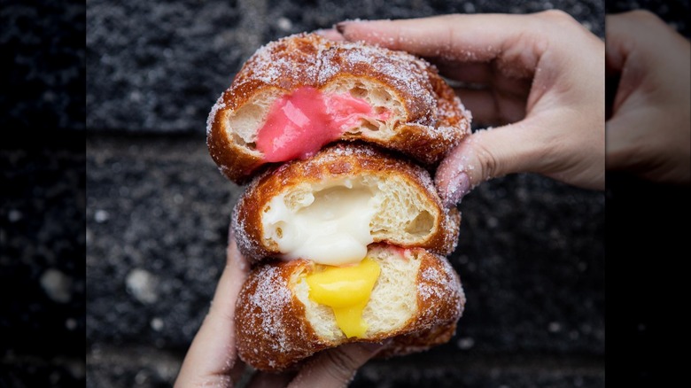 Hands holding a stack of three malasadas, each with a bite taken out to show a different fruit filling: guava, haupia, mango