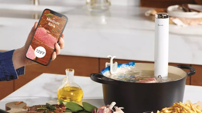 Breville Joule Turbo Sous Vide cooking a steak on the countertop with various ingredients and a hand holding a smartphone with an image of a medium rare steak on it