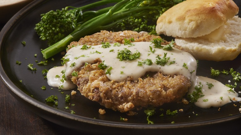 Chicken fried steak with gravy, broccolini, and a buttered roll on a black plate