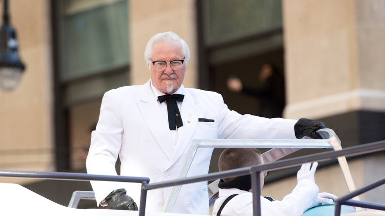 Colonel Sanders is standing on a parade float, in his iconic white suit, black bow tie and black gloves.