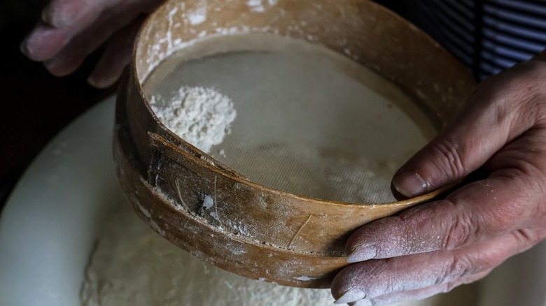 Close-up of a person sifting flour through a traditional round wooden sifter into a bowl.