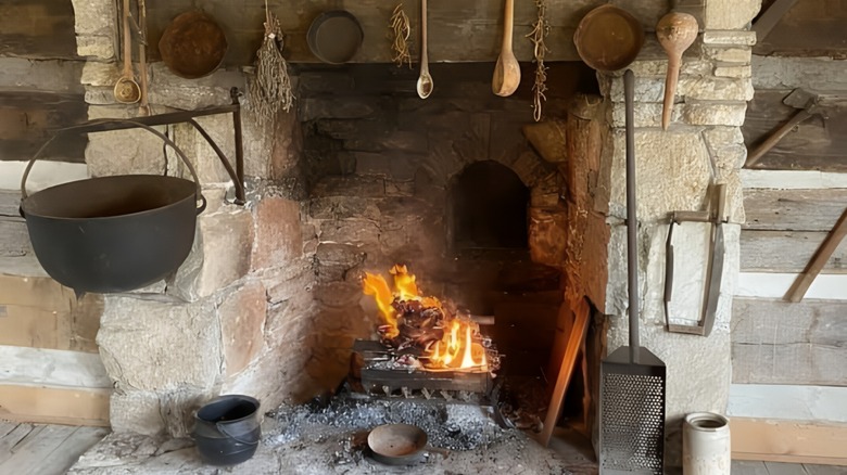 A rustic stone fireplace in a historic log cabin, featuring antique cookware, wooden utensils, and a glowing fire. A cast iron pot hangs over the flames, evoking the charm of pioneer-era cooking.