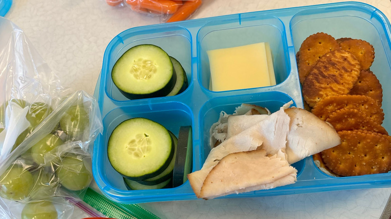 Adult packed lunch with veggies, cheese, crackers, and meats next to bags of grapes and carrots