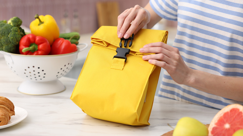 A woman packing a yellow lunch bag on a kitchen counter, surrounded by bowls of fruits and vegetables