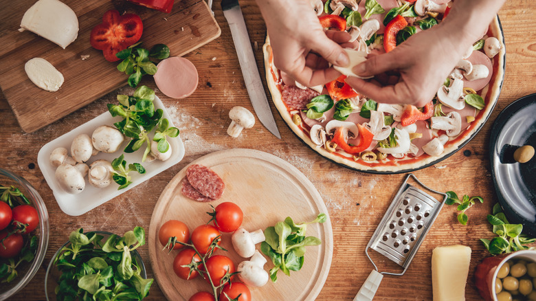 A pizza surrounded by many different ingredients. Hands are placing some ingredients on the pizza.