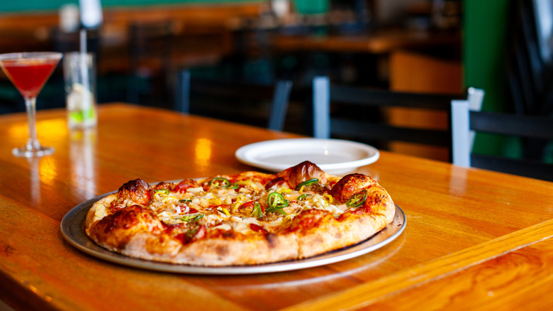A pizza rests on a metal try on a table inside a restaurant.