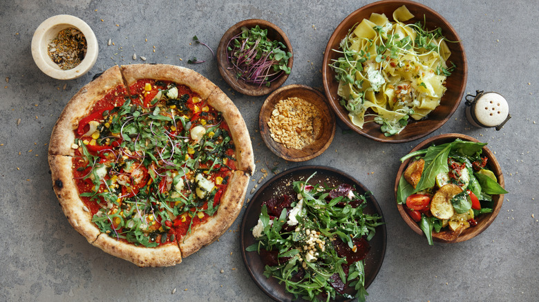 A pizza surrounded by wooden dishes of pasta, different types of salads and some garnish options.