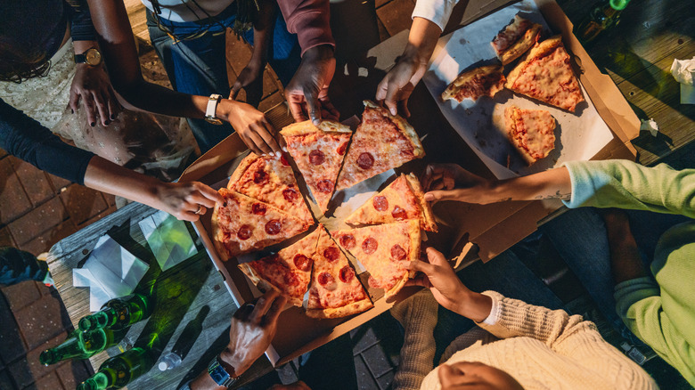A pizza sits in the middle of a pizza box on a table. It is surrounded by people who are pulling slices out of the box.