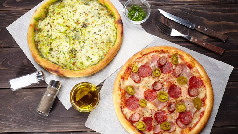 Two uncut pizzas rest on squares of parchment paper paper surrounded by cutlery, garnishes, and salt and pepper shakers.