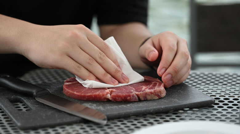 Close-up of a person's hands patting raw beef dry with paper towel
