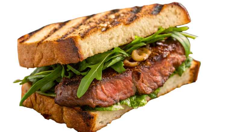 A steak sandwich on grilled bread against a white background